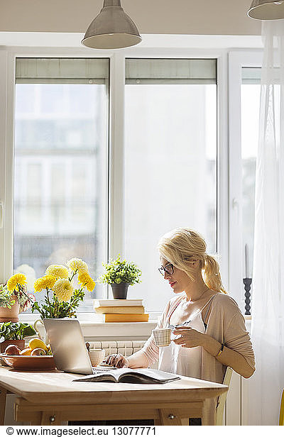 Woman with drink using laptop computer while sitting by window at home