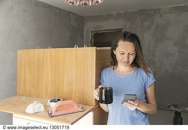 Woman with drink using phone while standing by furniture at home during renovation