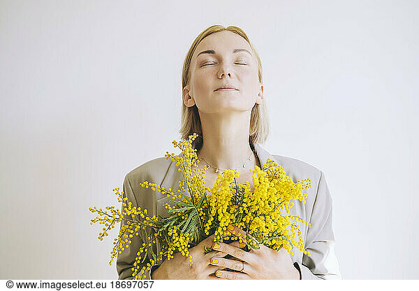 Woman with eyes closed holding mimosa flowers against white background
