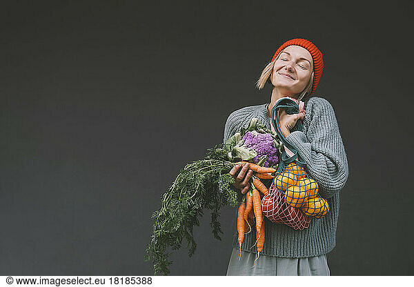 Woman with eyes closed holding organic fruits and vegetables in front of gray wall