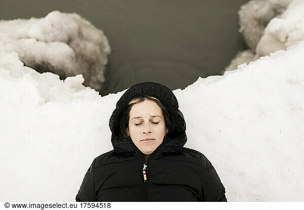 Woman with eyes closed lying down on snow by lake in winter