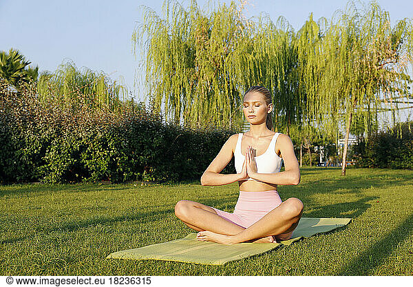 Woman with eyes closed practicing padmasana pose on exercise mat in park