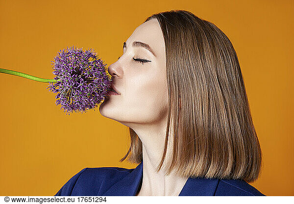Woman with eyes closed smelling allium flower against orange background