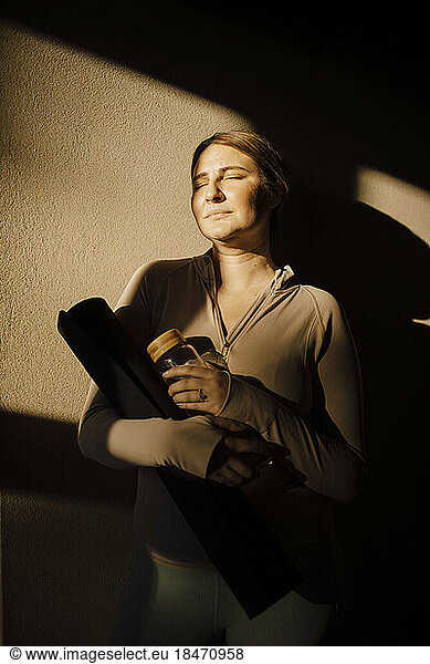 Woman with eyes closed standing against wall with sunlight on face