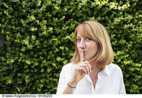 Woman with finger on lips standing in front of green hedge