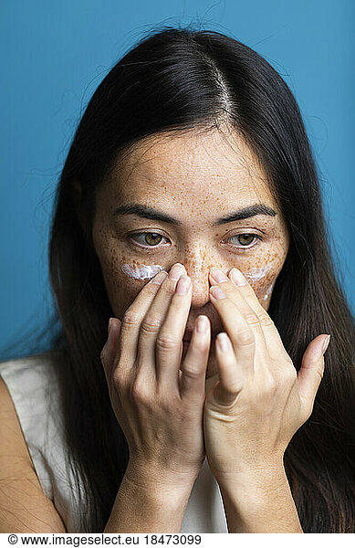 Woman with freckles applying lotion on face against blue background