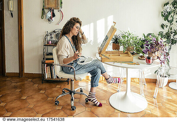 Woman with hand in hair sitting on chair painting in living room at home