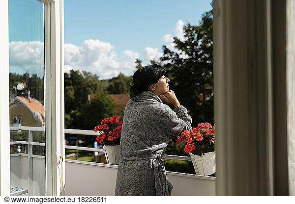 Woman with hand on chin enjoying sunlight while leaning on railing in balcony
