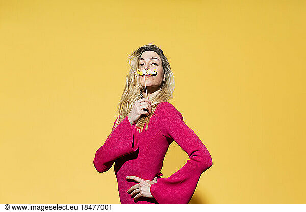 Woman with hand on hip holding fake mustache against yellow background