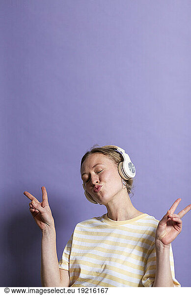 Woman with headphones gesturing peace sign against purple background