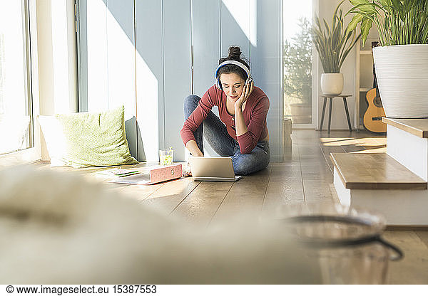Woman with headphones sitting at the window at home using laptop