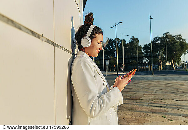 Woman with headphones using mobile phone leaning on wall