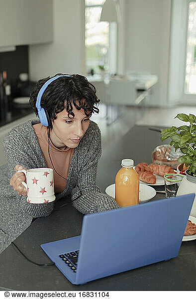 Woman with headphones working from home at laptop in morning kitchen