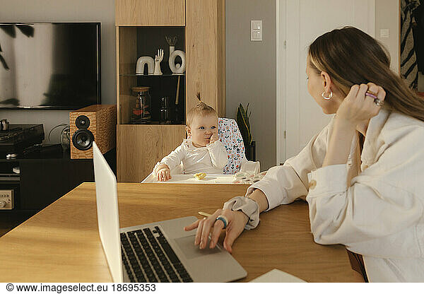 Woman with laptop looking at baby girl sitting in high chair at dining table