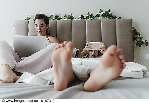 Woman with laptop sitting by girl using smart phone lying on bed at home