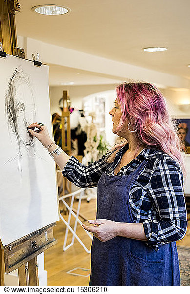 Woman with long blond wavy hair with pink streaks wearing apron standing at an easel  drawing a portrait.