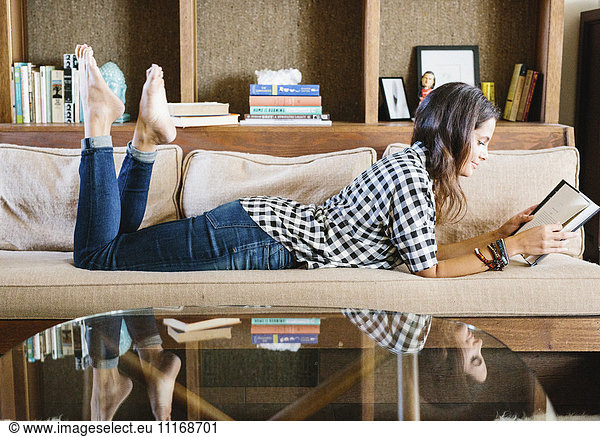 Woman with long brown hair lying on a sofa  reading a book.