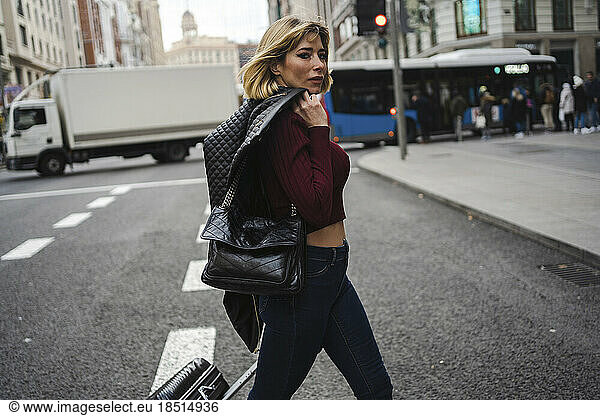 Woman with luggage and leather jacket crossing road in city