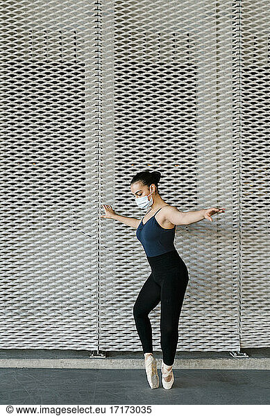 Woman with protective face mask practicing ballet dance by metal wall