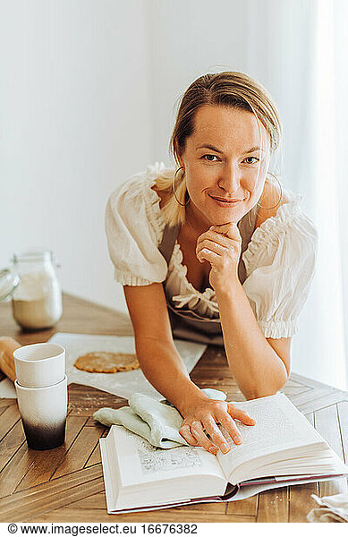 Woman with recipe book looking at camera while cooking at kitchen and