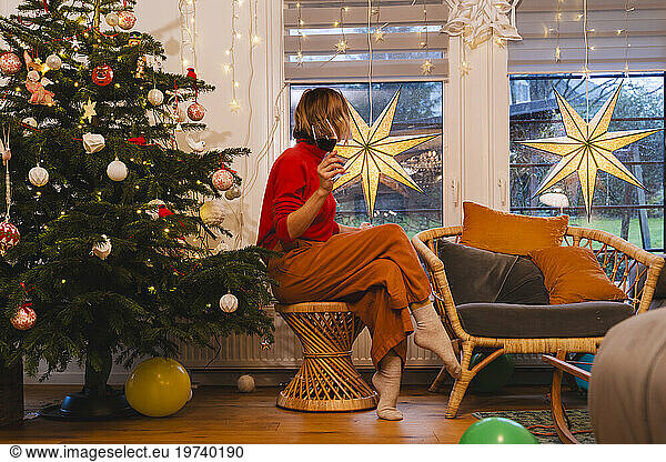 Woman with red wineglass looking through window at home during Christmas