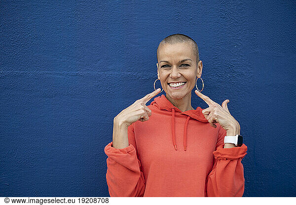 Woman with shaved head pointing and smiling in front of blue wall