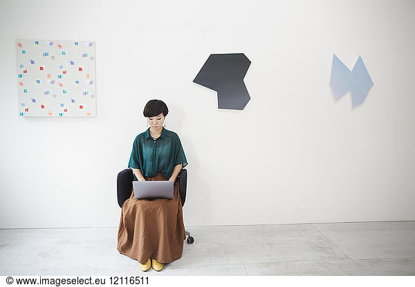 Woman with short black hair wearing green shirt sitting in art gallery  typing on laptop computer.
