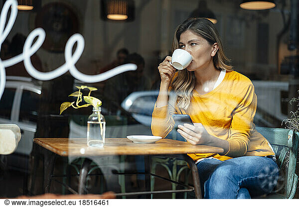 Woman with smart phone having coffee in cafe seen through glass