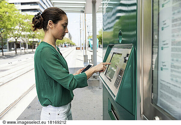 Woman with smart phone using ticket machine at tram stop