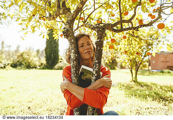 Woman with tablet PC hugging orange fruit tree in garden