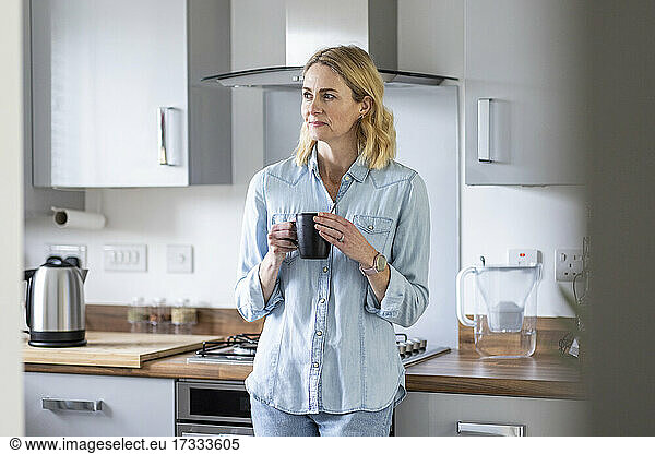 Woman with tea cup looking away while standing in kitchen at home
