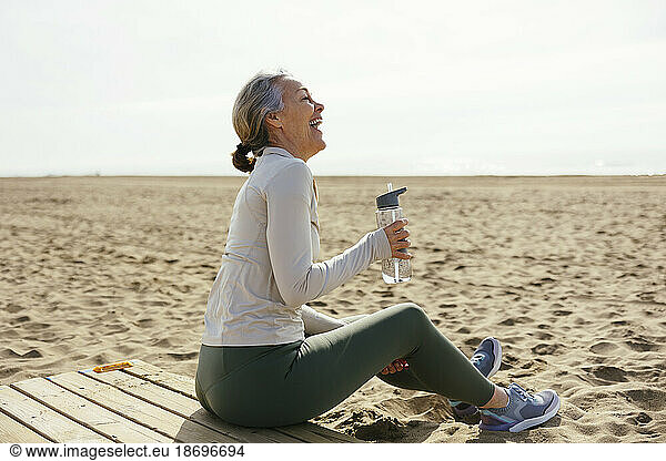 Woman with water bottle sitting and laughing at beach