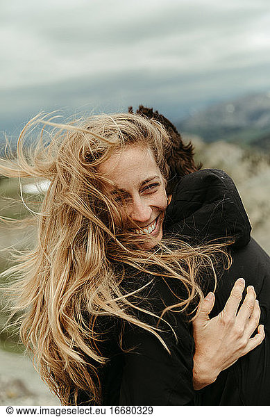 woman with wind swept long blonde hair laughs and hugs her partner