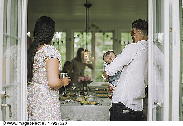 Woman with wineglass standing by man carrying daughter at party seen through doorway