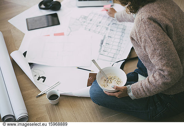 Woman working at home sitting on the floor looking at blueprint having breakfast
