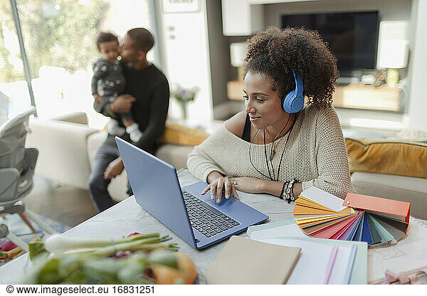 Woman working at laptop with husband and baby daughter in background