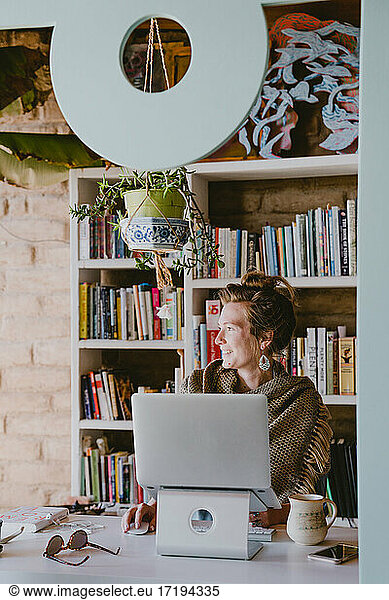 Woman works on laptop from home office surrounded by books and plants