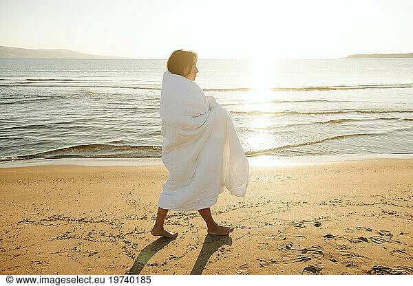 Woman wrapped in blanket walking barefoot on sand at beach