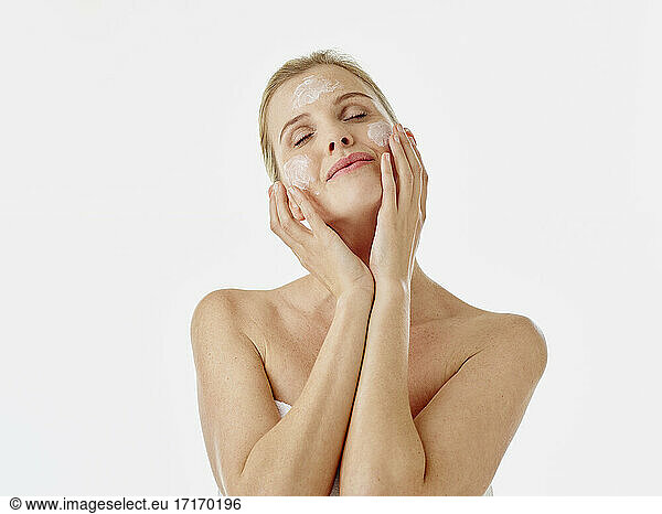 Woman wrapped in towel applying face cream while standing against white background