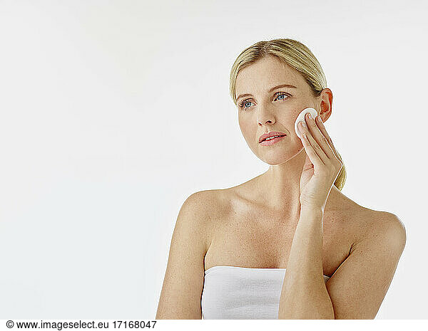 Woman wrapped in towel using cotton pad while standing white background