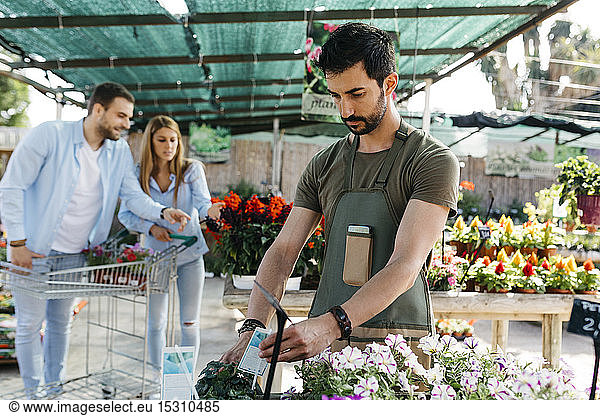 Worker in a garden center placing labels on flowers with customers in background