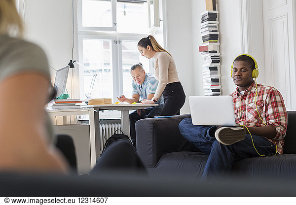 Worker using laptop on sofa and listening music  colleagues working in background