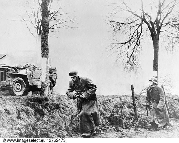 WORLD WAR II: COMBAT  1944. German soldiers file past a burning American half-track. Photographed 17 December 1944.