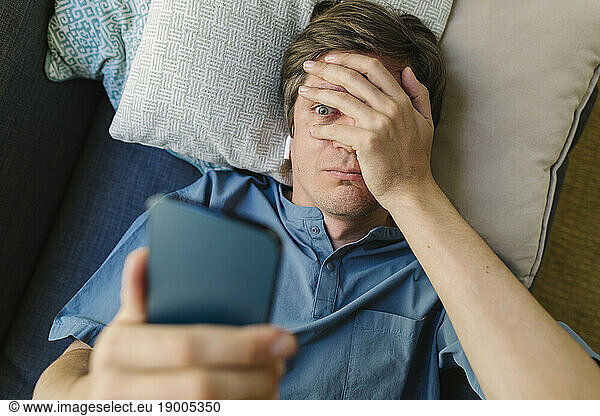 Worried man looking at smart phone lying on bed