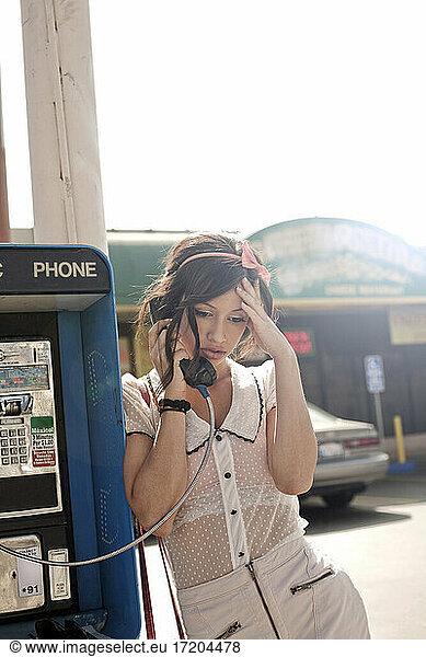 Worried woman with head in hand talking on pay phone at street during sunny day