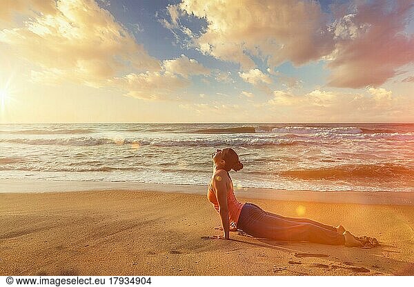 Yoga im Freien am Strand  Frau praktiziert Ashtanga Vinyasa Yoga Surya Namaskar Sun Salutation asana Urdhva Mukha Svanasana  nach oben gerichtete Hundestellung bei Sonnenuntergang. Mit Streulicht und Linsenreflexion