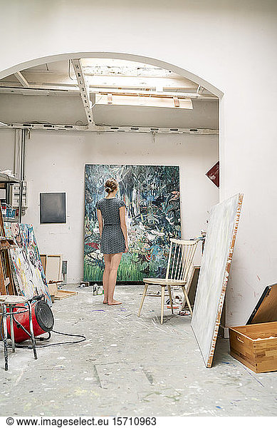 Young artist standing in her studio  looking at painting  rear view