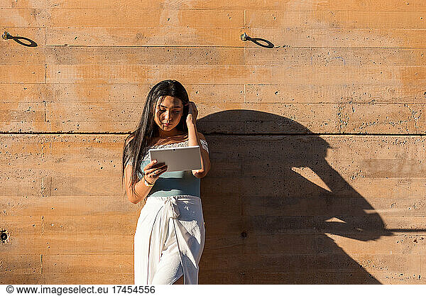 Young Asian girl leaning against a wall using her tablet.