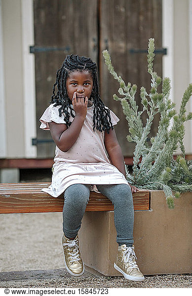 Young black girl with fingers in her mouth sitting on wood bench