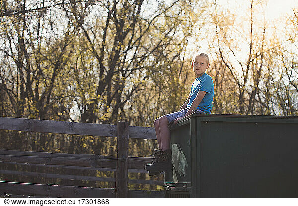 Young blond boy sitting outdoors by a fence at sunset.